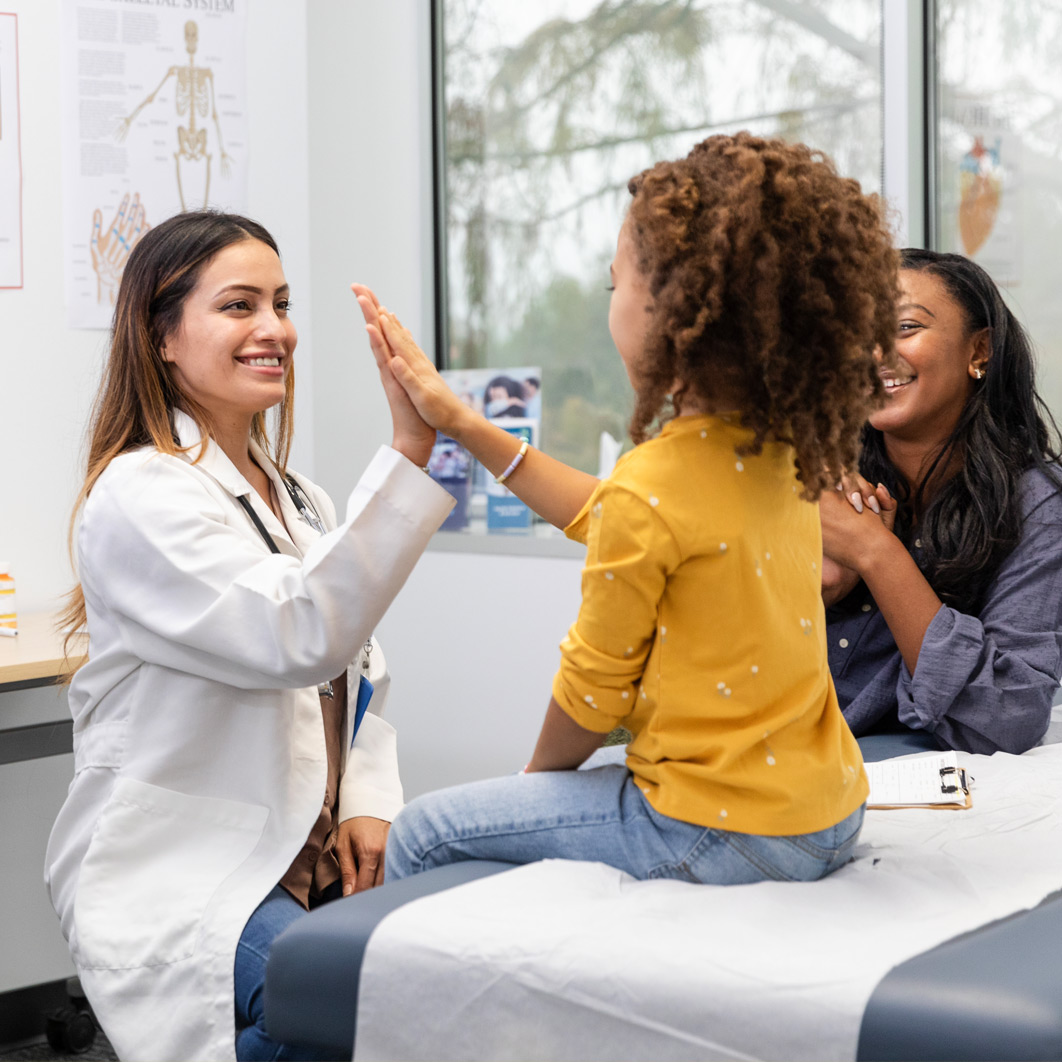 Girl giving a female doctor a high five while sitting on an examination table with her mother by her side